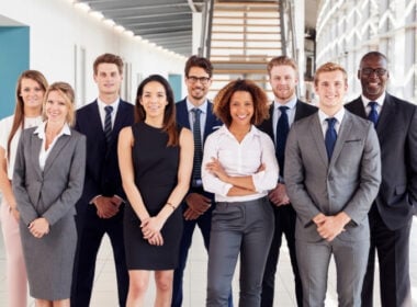 A group of nine professionally dressed people stand indoors, smiling at the camera. They are in a modern office building with glass walls and a staircase in the background.