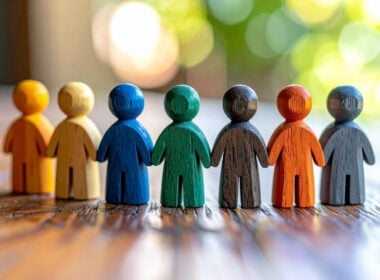 Seven colorful wooden human figures stand in a row on a wooden surface, with a blurred background of green and yellow light.