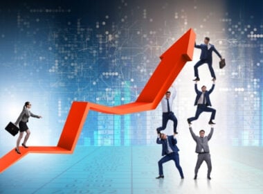 A businesswoman walks up a rising red arrow, while four businessmen in suits form a human pyramid, helping lift another man who stands at the top, all against a backdrop of financial graphs and digital data.