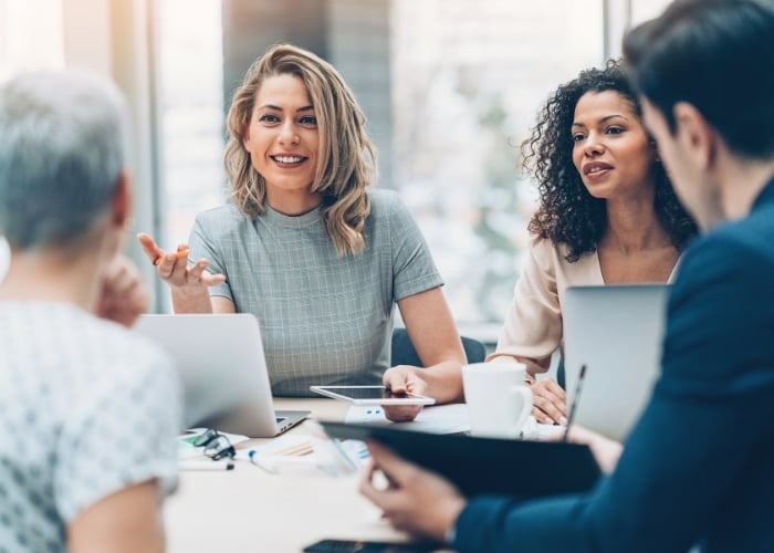 Four people sit around a table in a modern office, engaged in discussion. Two women face the camera, smiling and talking, with laptops and notebooks in front of them. The atmosphere is collaborative and professional.