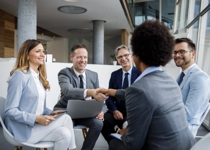 Five Professionally Dressed People Are Seated in a Modern Office Smiling and Interacting Two of Them Are Shaking Hands Suggesting a Successful Business Meeting or Agreement