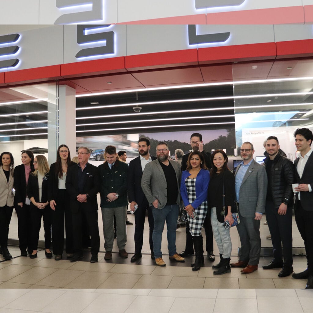 A group of fourteen people dressed in business attire stand in a line inside a modern building with bright lights and a large sign overhead.