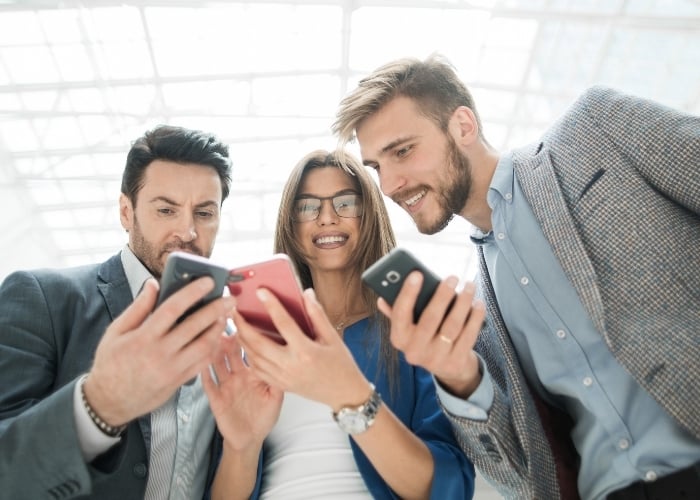 Three Young Professionals Two Men and One Woman Smiling and Looking at Their Smartphones Together in a Bright Modern Indoor Setting