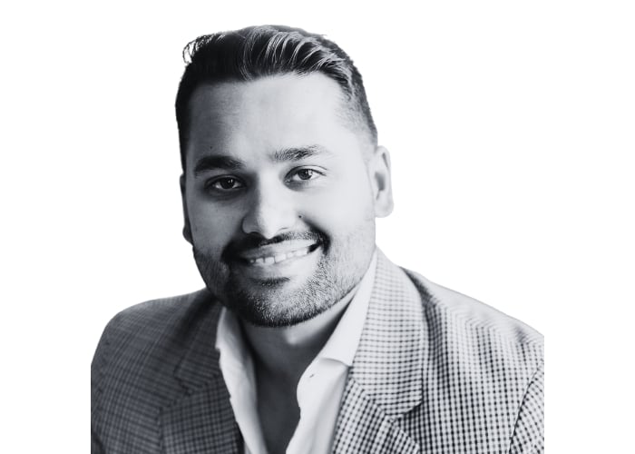 A man with short, styled hair and a trimmed beard, wearing a checkered blazer and a white shirt, smiles at the camera. The image is in black and white with a plain white background.