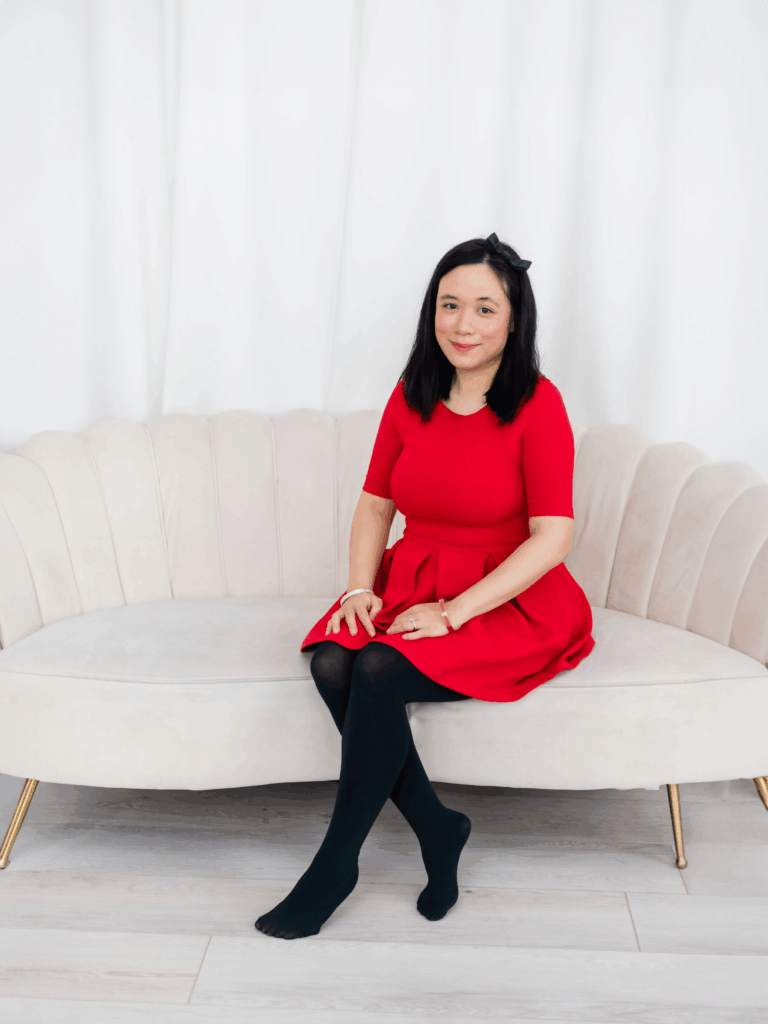 a Woman with Straight Black Hair Wearing a Red Dress and Black Tights Sits Barefoot on a Light colored Modern Sofa Against a White Background Smiling Slightly at the Camera