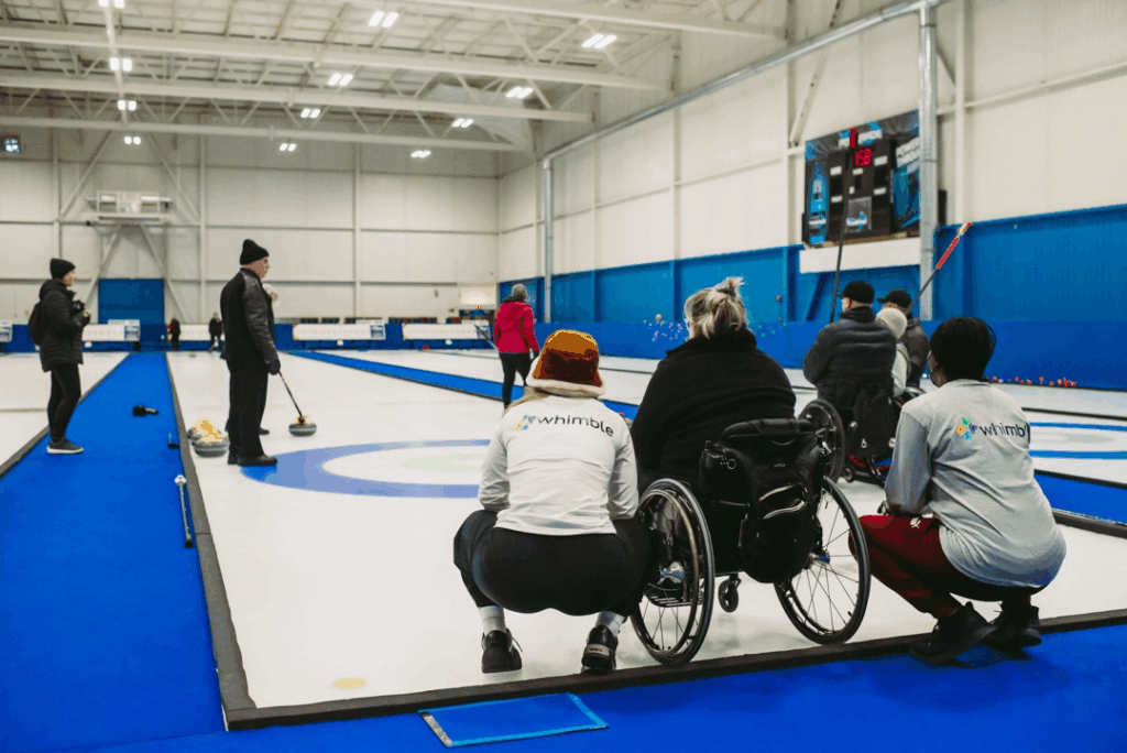 a Group of People Including a Person in a Wheelchair Participate in a Curling Game on an Indoor Ice Rink Some Players Wear Shirts with the Whimble Logo and Watch the Curling Stones on the Ice