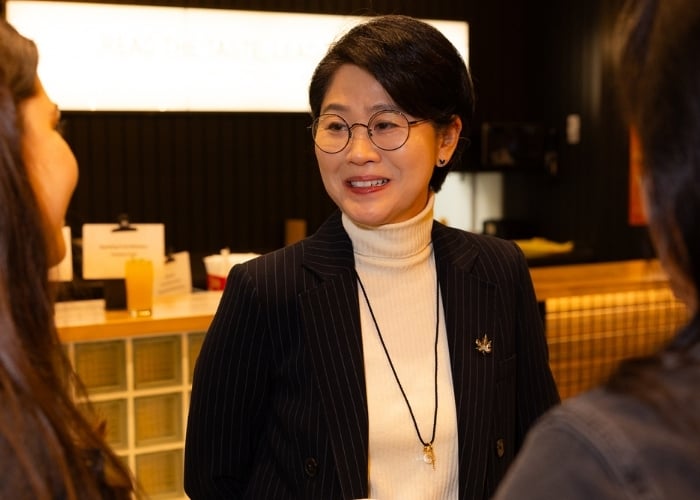 A woman with short black hair, glasses, and a white turtleneck under a dark pinstripe blazer smiles while talking to two people indoors. The background is warmly lit, with a counter and a blurred sign visible.