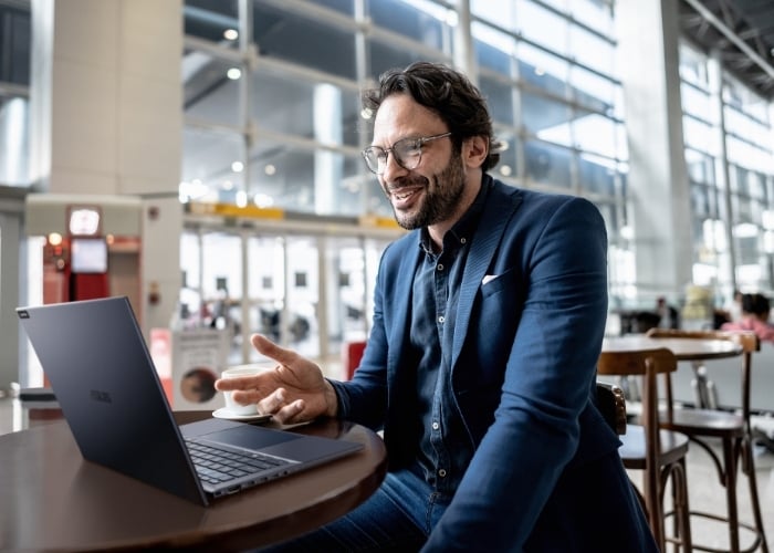 A man wearing glasses and a blue blazer sits at a table in a bright, modern airport terminal, smiling and gesturing while having a video call on his laptop. A cup of coffee is on the table beside him.
