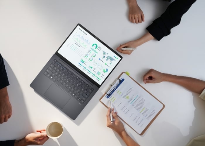 Three people sit at a white table with a laptop showing data charts, a clipboard with documents, and a coffee cup, appearing to be in a business meeting or discussion.