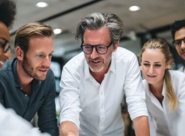 A group of five people, three men and two women, gather closely around a table, engaged in discussion and smiling, in a modern office setting.