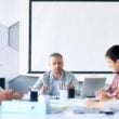 a Group of Six People Sit Around a Conference Table Having a Discussion One Man in the Center is Speaking While Others Listen Attentively There Are Notebooks Pens and a Laptop on the Table and a Blank Screen in the Background