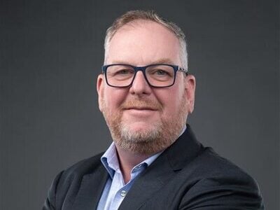 A man with short hair, a beard, and glasses wearing a dark suit jacket and light blue shirt stands with his arms crossed, smiling at the camera against a plain dark background.
