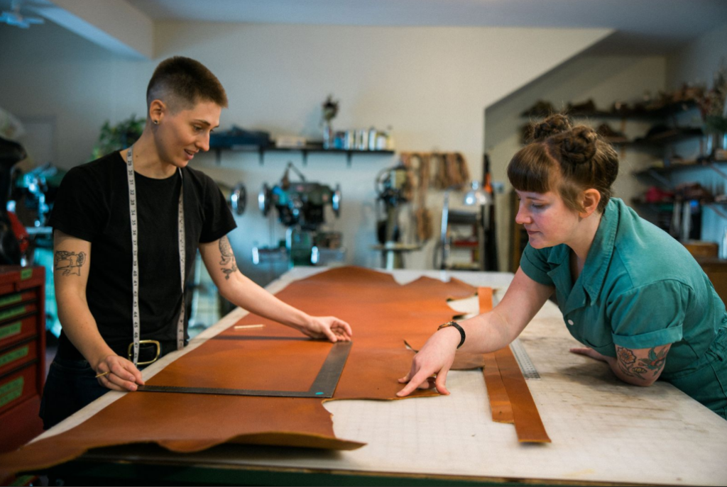 Two people work together in a workshop, measuring and cutting large pieces of brown leather on a table. One wears a black shirt and holds a tape measure; the other, in green, gestures at the leather, both focused on the task.