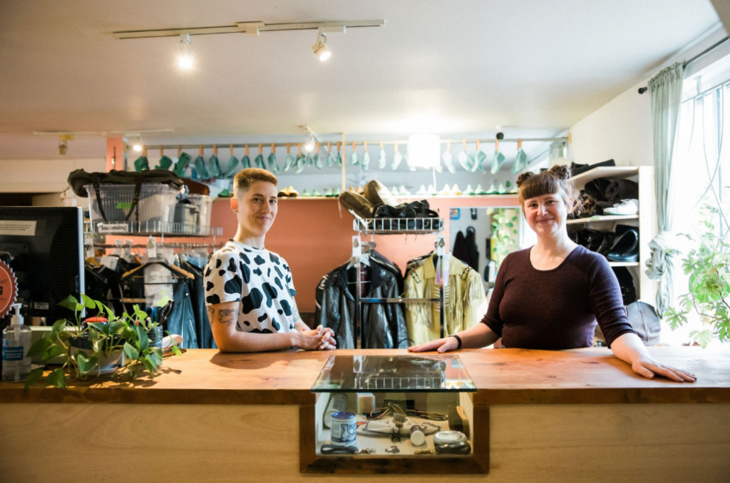 Two people stand behind a wooden counter in a well-lit shop, surrounded by clothing, plants, and sewing supplies. Both are smiling, and the shop has a welcoming, creative atmosphere.