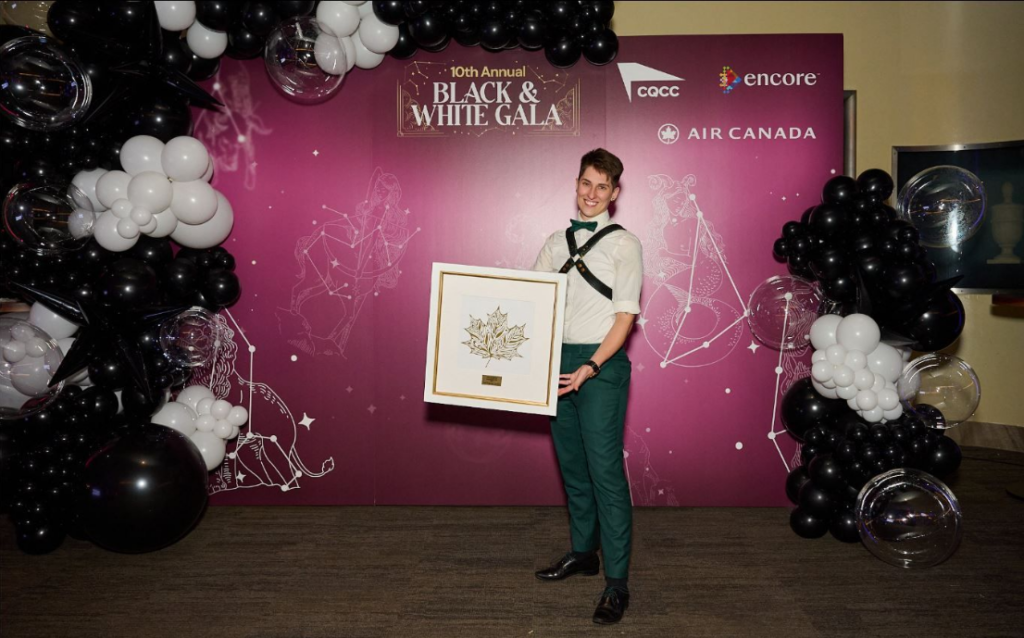 A smiling man in green pants and suspenders holds a framed artwork in front of a purple backdrop that reads 10th Annual Black & White Gala, surrounded by black and white balloon decorations.