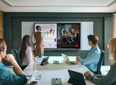 A man stands at a digital whiteboard presenting to four colleagues seated at a conference table, while a video call with a group of people is displayed on a large screen beside him. Laptops and tablets are on the table.