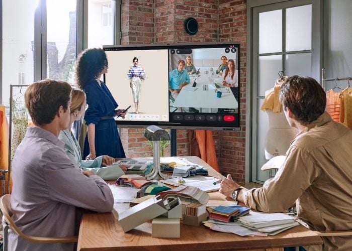 Four people sit around a table with fabrics, watching a video conference on a large screen. One screen shows a fashion model, the other shows a group of people in a meeting. The setting is a modern workspace.