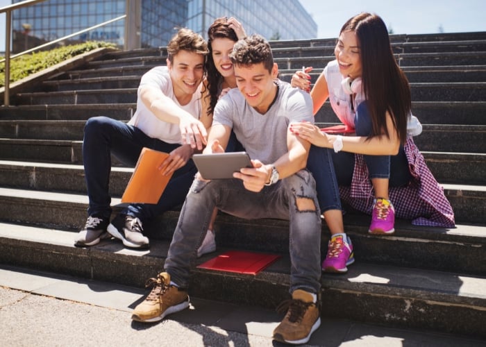 Four smiling young adults sit on outdoor steps, looking at a tablet together. Two hold folders, and they appear to be enjoying each others company in a casual, sunny setting.