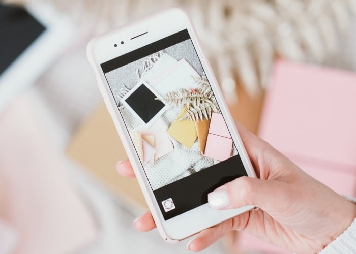 A hand holds a smartphone displaying a photo of scattered pastel envelopes, polaroid photos, and a small plant on a textured surface. The background features blurred pink and beige objects.