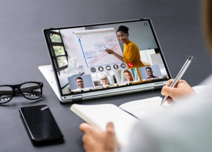 A person takes notes while attending a virtual meeting on a tablet, where a woman gives a presentation on a flip chart and four colleagues participate via video call.