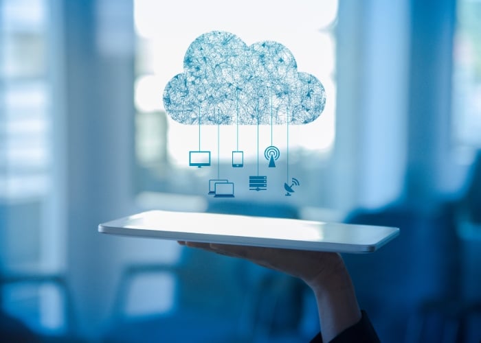 A hand holds a tablet with a digital cloud above it, connected to icons representing various devices and technologies, symbolizing cloud computing and data connectivity. The background is blurred and blue-toned.