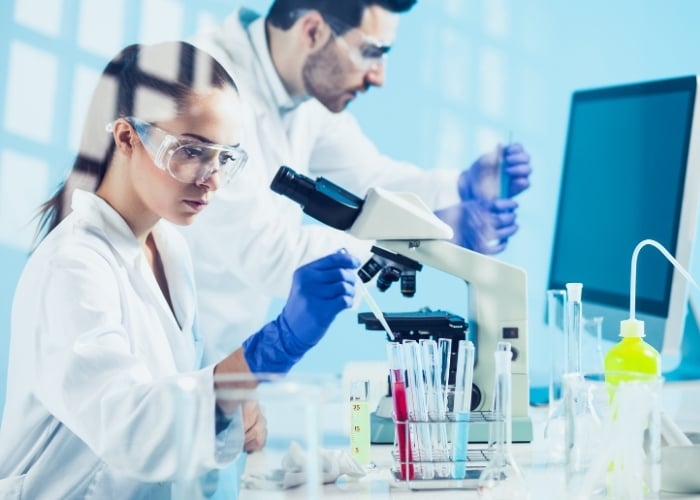 Two scientists in lab coats and safety goggles work in a laboratory; one examines samples with a pipette near a microscope, while the other works in the background, with test tubes and lab equipment on the desk.