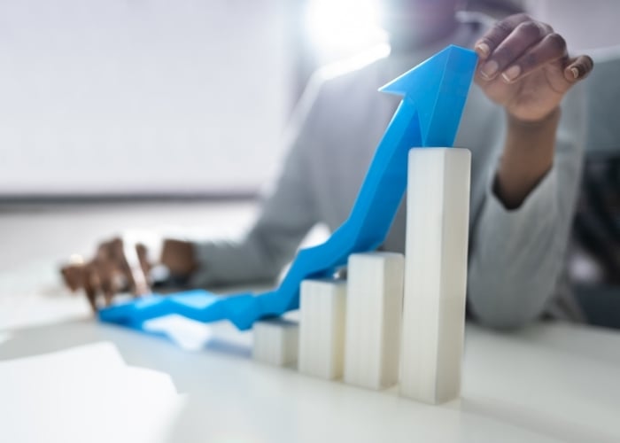 A person’s hand adjusts a blue upward arrow resting on white bar graph columns, symbolizing growth or business success, with soft lighting and a blurred background.