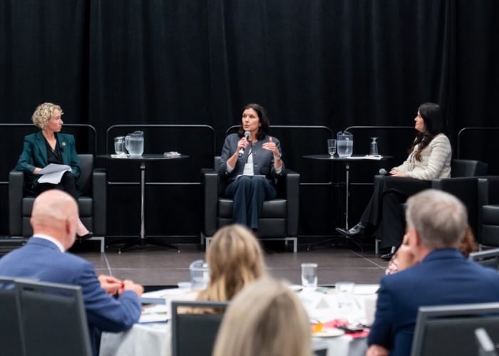 Three women sit on stage in armchairs during a panel discussion; one woman speaks into a microphone while the others listen. Audience members are seated in the foreground. Black curtain backdrop and water pitchers are visible.
