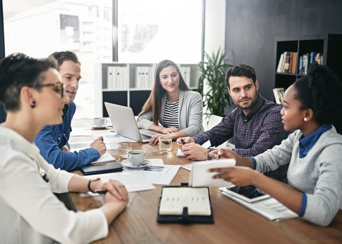 Five people sit around a table in a modern office, engaged in discussion. Papers, a notebook, a tablet, and a laptop are on the table. The group appears to be collaborating on a project or having a meeting.