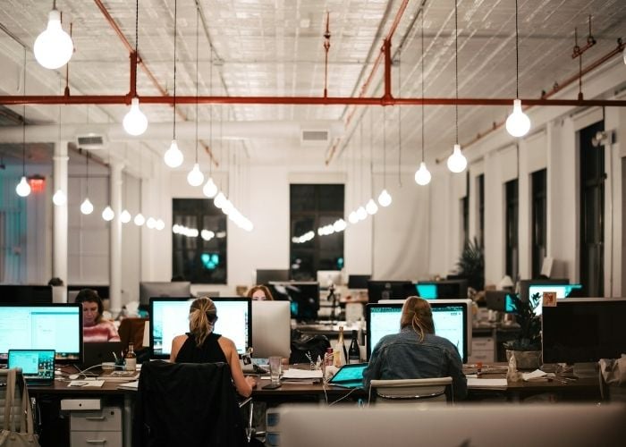 A modern open-plan office with several people working at desks with computers, under rows of hanging light bulbs. The workspace has a bright, collaborative atmosphere.