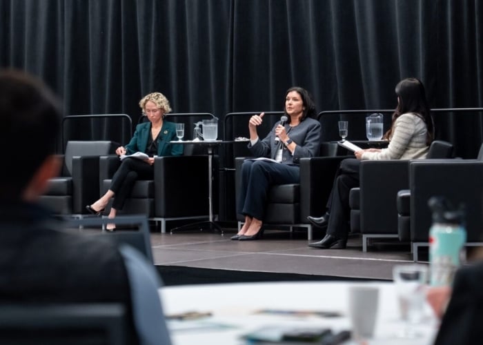 Three women sit on a stage in armchairs, engaged in a panel discussion. One woman speaks into a microphone while the others listen and take notes. A black curtain serves as the backdrop.