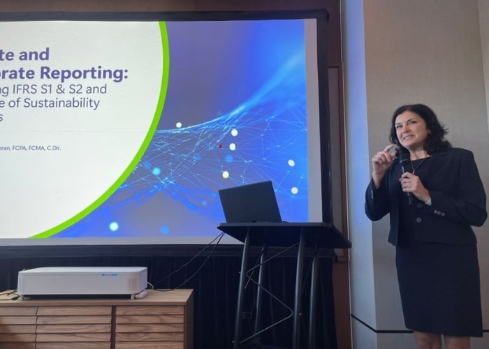 A woman in business attire speaks into a microphone while standing next to a podium. Behind her is a presentation slide about climate and corporate reporting, referencing IFRS S1 & S2 and sustainability.