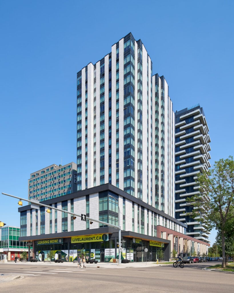 A tall modern apartment building with vertical white and gray panels, large windows, and commercial shops at street level stands on a city corner. People walk nearby and a cyclist passes on the road.