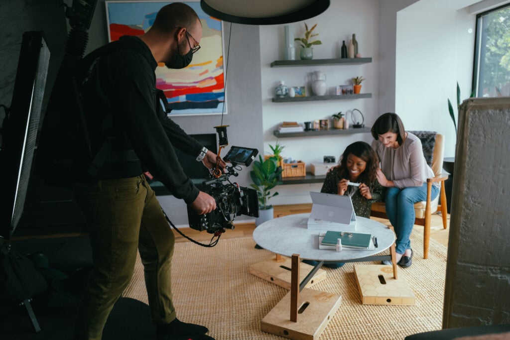 A cameraman films two women sitting at a round table working on a laptop in a brightly lit, modern living room with shelves, plants, and a large colorful painting on the wall.