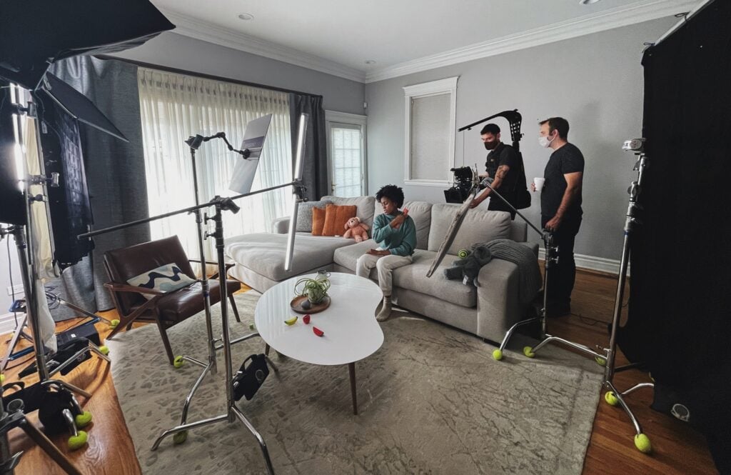 A film crew sets up lights and cameras around a boy sitting on a living room couch with a stuffed animal, while two crew members in masks stand behind the equipment.