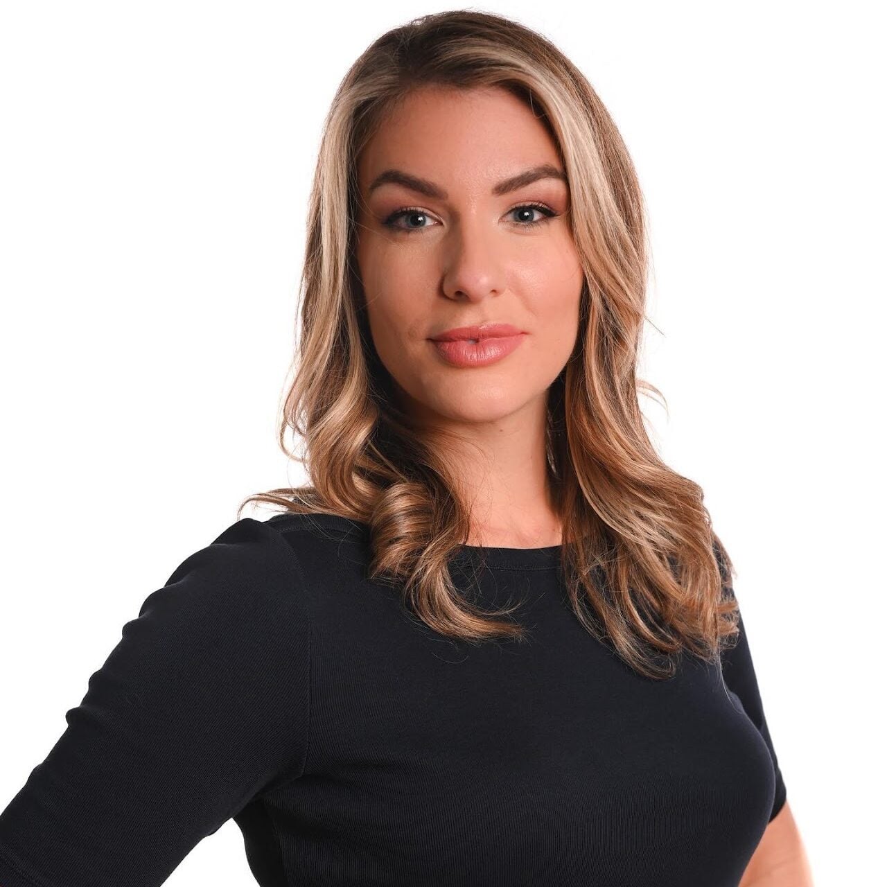 A woman with wavy, shoulder-length blonde hair wearing a black short-sleeve top stands confidently against a white background, looking directly at the camera with a slight smile.