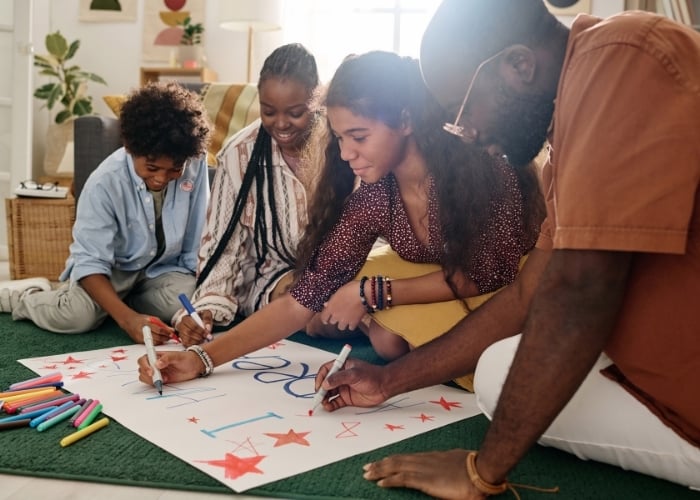 Four people sit on the floor together, smiling and drawing colorful designs with markers on a large poster. The poster features stars, letters, and bright colors. Art supplies are scattered nearby.
