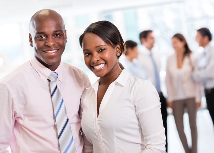 Two professionals, a man and a woman, stand smiling in the foreground, dressed in business attire. In the blurred background, several colleagues are talking in an office setting.