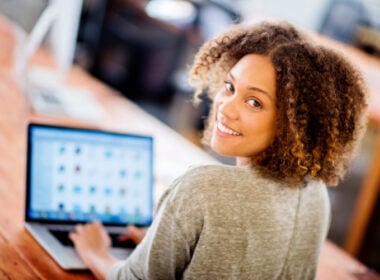 A person with curly hair smiles at the camera while sitting at a wooden table and using a laptop computer, with blurred office surroundings in the background.