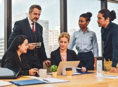 Five business professionals, three women and two men, gather around a desk in an office with large windows, discussing work as one woman types on a laptop. Papers, coffee cups, and a small plant are on the table.