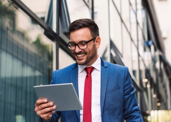 A man in a blue suit and red tie smiles while holding and looking at a tablet outdoors, with modern glass buildings reflected in the background.