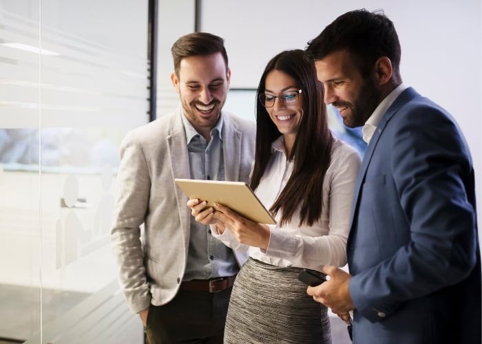 Three professionally dressed people stand together in an office, smiling and looking at a digital tablet held by the woman in the center. The atmosphere appears collaborative and positive.