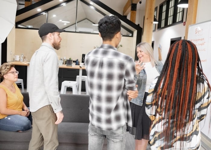 Five people gather in a modern office space, some standing and some sitting. One woman, smiling and gesturing, leads the conversation. A whiteboard and kitchen area are visible in the background.