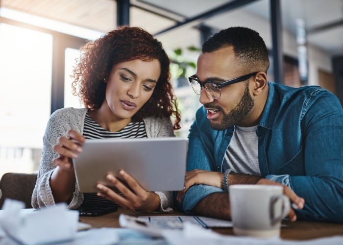 A woman and man sit together at a table, looking at a tablet. Papers and a coffee mug are on the table in front of them, and they appear focused and engaged in conversation.