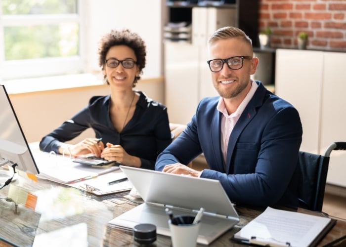 Two business professionals, a woman and a man, sit at a desk in a modern office, smiling at the camera. Laptops, documents, and office supplies are visible on the desk in front of them.