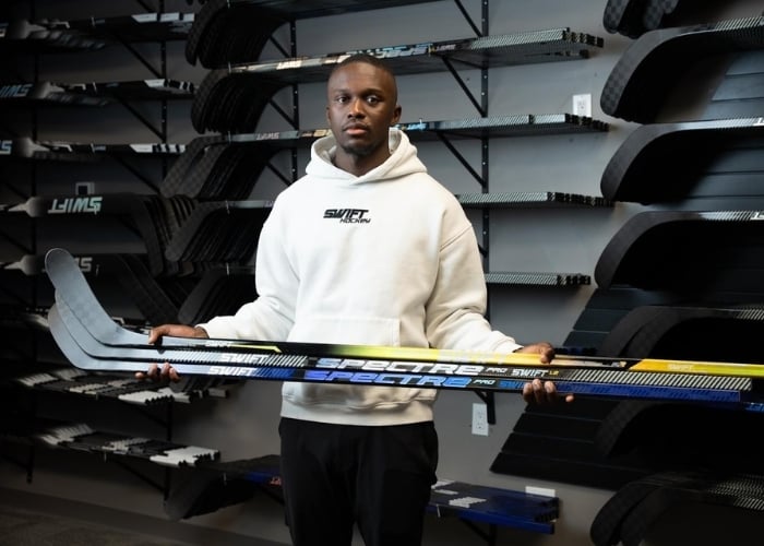 A man in a white hoodie holds two hockey sticks inside a store, standing in front of a wall displaying multiple hockey sticks.