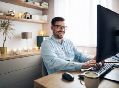 A smiling man with glasses sits at a desk in a home office, using a computer. Shelves with books, decorations, and a plant are in the background, and a mug is next to him on the desk.