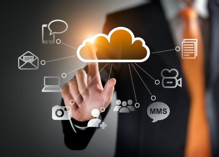A person in a suit touches a virtual cloud icon, with various technology and communication icons like email, video, mobile, and chat connected around it, symbolizing cloud computing and data connectivity.
