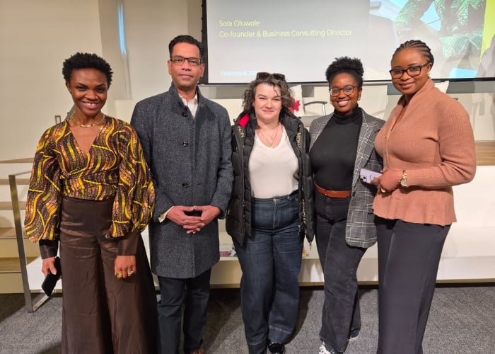 Five people stand together indoors, smiling at the camera. Behind them is a presentation slide displaying “Sola Olukowle, Co-founder & Business Consulting Director.” The group is dressed in smart, business-casual attire.