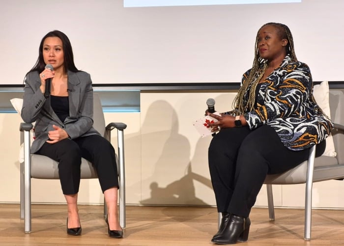 Two women sit on stage in chairs, each holding a microphone. The woman on the left wears a gray blazer and black pants; the woman on the right wears a patterned jacket and black pants. They appear to be speaking at an event.